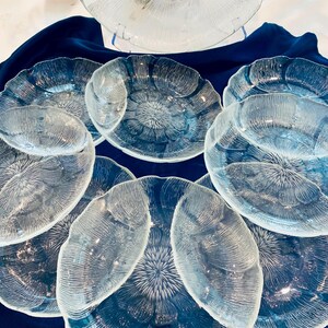 May include: A collection of clear glass serving dishes with a floral design. The set includes several small plates and a large serving bowl. The dishes are arranged on a dark blue cloth, creating a contrast that highlights their intricate patterns.