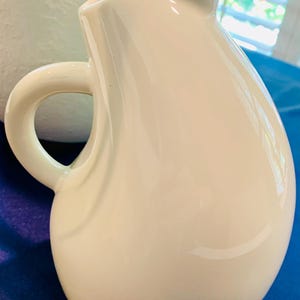 May include: A glossy, off-white ceramic pitcher with a smooth, rounded, teardrop-shaped body. The pitcher features a curved handle on the left side and a pointed pouring spout at the top, showcasing a clean, solid-colored, reflective surface.