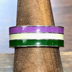Peut inclure: Une bague en argent avec trois bandes de couleurs : violet, blanc cassé et vert. La bague est présentée sur un présentoir en bois. Les couleurs sont vives et la bague a une finition polie.