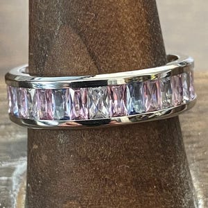 Puede incluir: Un anillo plateado con una banda de piedras rectangulares rosas y celeste claro. El anillo se exhibe en un soporte de madera. Las piedras están muy juntas, creando una banda continua de color alrededor del anillo.