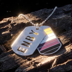 Puede incluir: Un collar con dos placas de identificación plateadas. La primera placa tiene la palabra "ENBY" recortada. La segunda placa tiene un diseño colorido con rayas amarillas, blancas, rosas, moradas, negras y grises. El collar está sobre un trozo de madera.