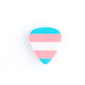 Può includere: Un plettro per chitarra di forma triangolare, con il design della bandiera del Pride Transgender. Il plettro ha una parte superiore e inferiore azzurra, con un centro a strisce rosa e bianche. Su sfondo bianco.