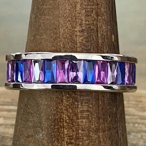 May include: A silver ring with a band of alternating blue and purple rectangular gemstones. The ring is displayed on a wooden ring holder, showcasing the vibrant colors and design of the jewelry.