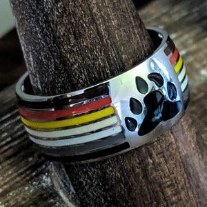 May include: A silver ring featuring a black paw print. The ring is decorated with bands of red, yellow, and white enamel. It is displayed on a wooden ring holder.