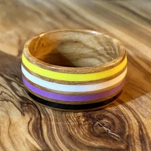 May include: A wooden ring with a non-binary pride flag design. The ring features horizontal stripes of yellow, white, purple, and black. The ring is made of light-colored wood and has a smooth finish.
