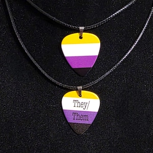 Può includere: Due collane a forma di plettro per chitarra. Quella superiore ha la parte superiore gialla, il centro bianco, la parte inferiore viola e la punta nera. La collana inferiore ha le parole "They/ Them". Entrambe sono su cordini neri.