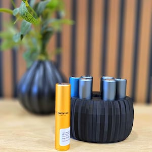 May include: An arrangement of perfume bottles in a black holder. One gold bottle stands out, with the brand name visible. Other bottles are blue and silver. A black vase with greenery is in the background, on a wooden surface.
