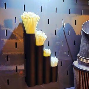 May include: A black four-tube organizer mounted on a gray pegboard, holding bundles of off-white plastic zip ties of varying lengths. The tubes are arranged in a stepped formation, with each section containing a different size of cable ties.