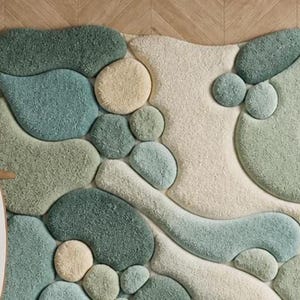 May include: A soft, abstract rug with a pebble design in green, blue, and cream. The rug has a textured, tufted surface, creating a soft feel. The organic shapes and muted colours give it a modern, natural aesthetic. The rug would look great in a living room.