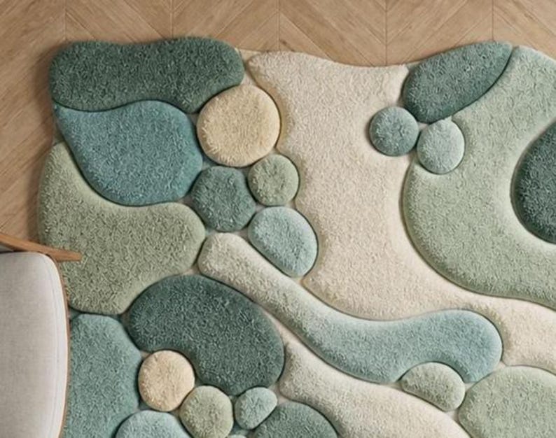 May include: A soft, abstract rug with a pebble design in green, blue, and cream. The rug has a textured, tufted surface, creating a soft feel. The organic shapes and muted colours give it a modern, natural aesthetic. The rug would look great in a living room.