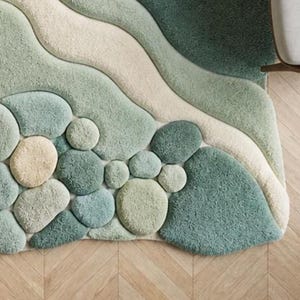 May include: A textured area rug with a pebble and wave design. The rug features light green, beige, and teal colours. The rug's design includes rounded shapes resembling stones and flowing wave patterns.