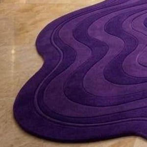 May include: A purple, wavy-shaped rug with a textured surface. The rug's design features multiple layers of varying shades of purple, creating a three-dimensional effect. The rug is placed on a light-colored, tiled floor.