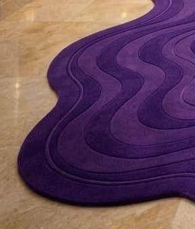 May include: A purple, wavy-shaped rug with a textured surface. The rug's design features multiple layers of varying shades of purple, creating a three-dimensional effect. The rug is placed on a light-colored, tiled floor.
