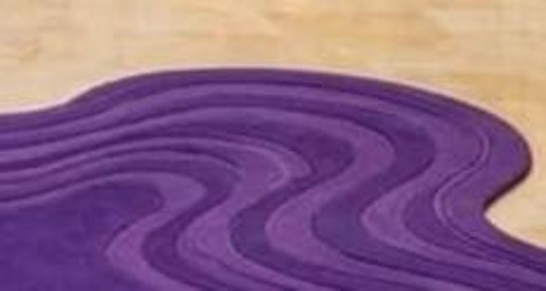 May include: A wavy, purple rug with a textured surface. The rug's design features a series of curved lines, creating a flowing, organic pattern. The rug is placed on a light-colored wooden floor.