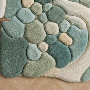 May include: A textured rug with a pebble design in shades of green, blue, and cream. The rug features a variety of rounded shapes, creating a three-dimensional effect. The rug is placed on a wooden floor.