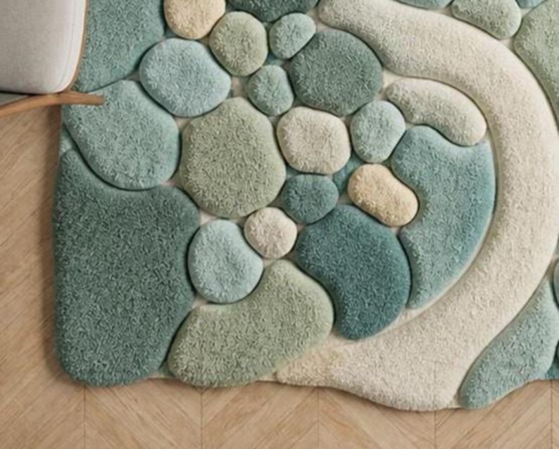 May include: A textured rug with a pebble design in shades of green, blue, and cream. The rug features a variety of rounded shapes, creating a three-dimensional effect. The rug is placed on a wooden floor.