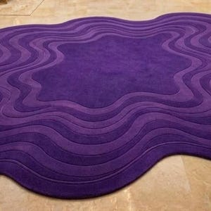 May include: A large, abstract rug in shades of purple. The rug has a unique, organic shape with layered, wavy patterns. The color palette ranges from deep violet to lighter shades, creating a textured, dimensional effect. The rug is placed on a light-colored tiled floor.