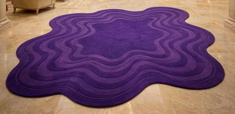 May include: A large, abstract rug in shades of purple. The rug has a unique, organic shape with layered, wavy patterns. The color palette ranges from deep violet to lighter shades, creating a textured, dimensional effect. The rug is placed on a light-colored tiled floor.