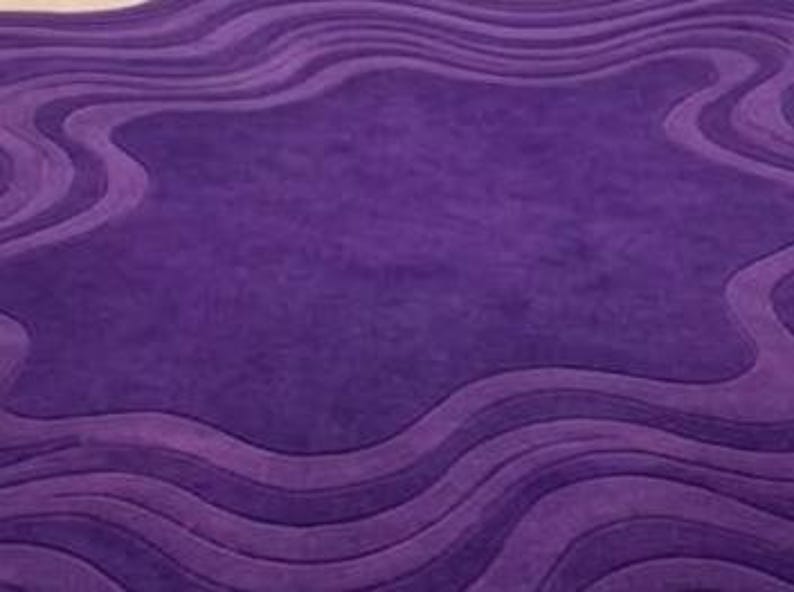 May include: A plush, purple area rug with a unique, wavy design. The rug features varying shades of purple, creating a textured, three-dimensional effect. The central area is a lighter shade, surrounded by darker, undulating borders.