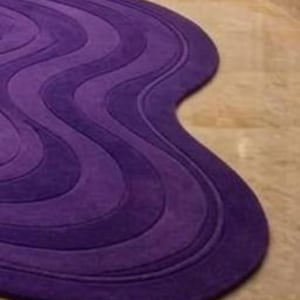 May include: A purple rug with a wavy design. The rug features varying shades of purple, creating a layered effect. The rug's shape is organic, with a flowing, non-rectangular outline. The rug is made of a soft material.