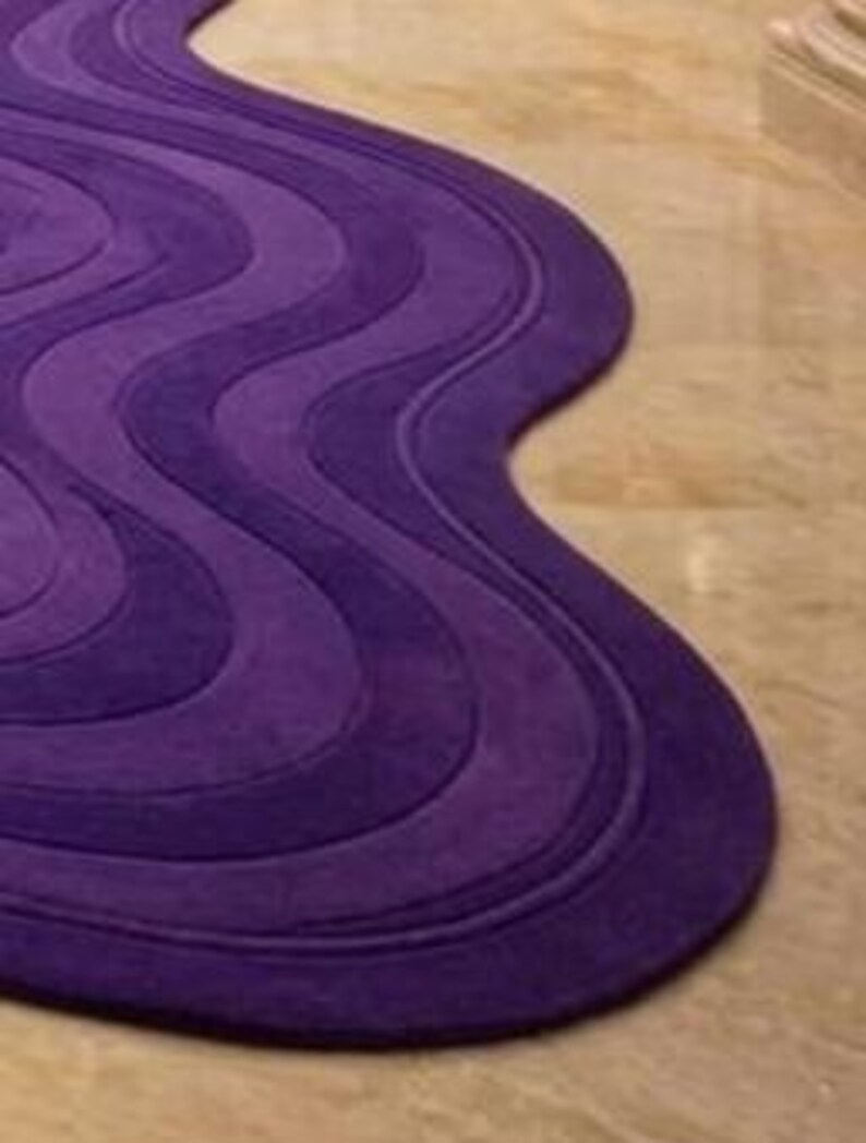 May include: A purple rug with a wavy design. The rug features varying shades of purple, creating a layered effect. The rug's shape is organic, with a flowing, non-rectangular outline. The rug is made of a soft material.