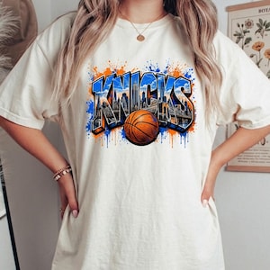 May include: A cream-colored short-sleeve t-shirt featuring a graphic print of the word KNICKS in blue and orange. The letters contain a detailed New York City skyline illustration, and an orange basketball is positioned in front of the text.