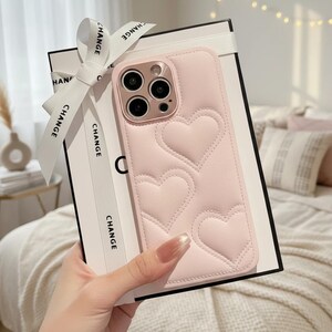 May include: A light pink phone case with a heart pattern. The case is on a black and white box with a white ribbon that says "CHANGE". The phone case has a camera and flash.