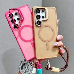 May include: Two phone cases: one pink and one tan. The pink case has a pink ring around the camera and a pink circle on the back. The tan case has a gold ring around the camera and a tan circle on the back. Both cases have a strap attached.