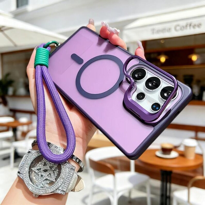 May include: A purple phone case with a matching camera lens protector and a purple lanyard. The case has a dark purple ring and a green and purple wrist strap. The phone case is held in a hand.