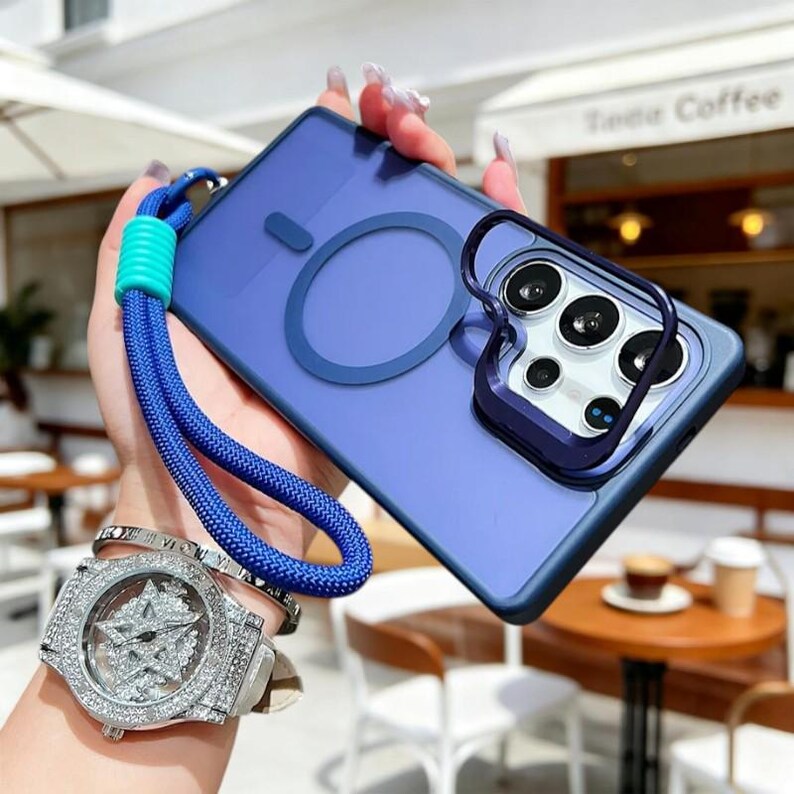 May include: A blue phone case with a matching blue lanyard. The case has a ring for a magnetic charger and a dark blue camera protector. The lanyard is blue with a turquoise accent. The phone case is held in a hand.