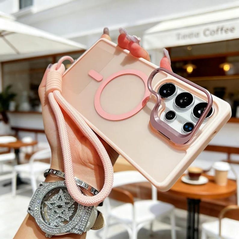 May include: A light pink phone case with a matching pink strap. The case has a circular design on the back and a metallic frame around the camera lenses. The case is held up by a pink strap.