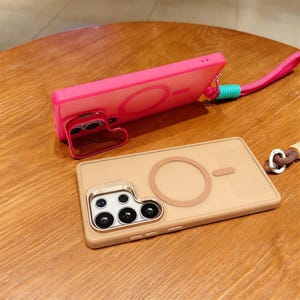 May include: Two phone cases, one pink and one tan, with a circular design and a small rectangular design. The pink case has a heart-shaped stand. Both cases have a lanyard attachment. The tan case has a camera module with multiple lenses.