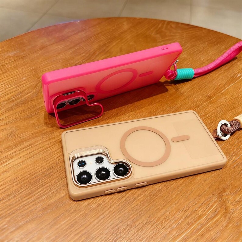 May include: Two phone cases, one pink and one tan, with a circular design and a small rectangular design. The pink case has a heart-shaped stand. Both cases have a lanyard attachment. The tan case has a camera module with multiple lenses.
