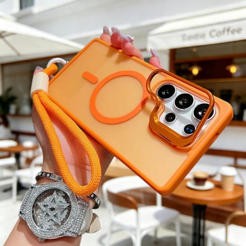 May include: Orange phone case with a matching orange strap. The case has a ring around the camera and a circular design on the back. The case is held by a white and orange strap.