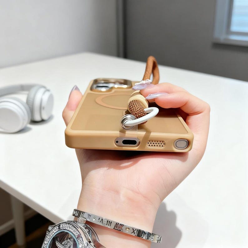 May include: A tan phone case with a brown strap and a white ring attachment. The case has a circular design on the back. A pair of white headphones are in the background.