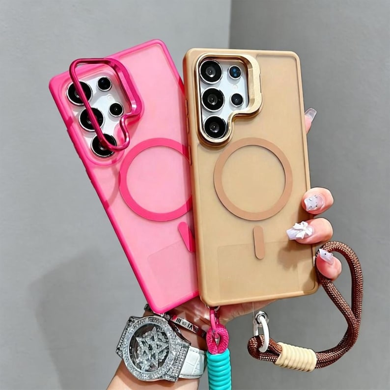 May include: Two phone cases: one pink and one tan. The pink case has a pink ring around the camera and a pink circle on the back. The tan case has a gold ring around the camera and a tan circle on the back. Both cases have a strap attached.