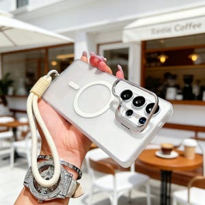 May include: Clear phone case with a white magnetic ring and a silver camera lens protector. The case has a cream-colored lanyard attached. The background is blurred, showing a coffee shop setting.