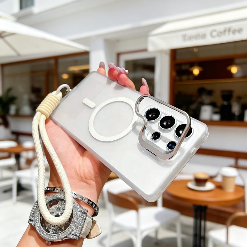 May include: Clear phone case with a white magnetic ring and a silver camera lens protector. The case has a cream-colored lanyard attached. The background is blurred, showing a coffee shop setting.