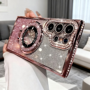 May include: A rose gold and silver glitter phone case with a ring holder and camera lens protector. The case is encrusted with rhinestones around the edges and camera lens. The ring holder has the text "KATHY TECH".