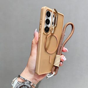 May include: A tan phone case with a gold ring holder and a matching brown lanyard. The case has a circular magnetic attachment and a vertical bar design. The lanyard features a woven cord and a series of beige beads. The phone case is held in a hand.