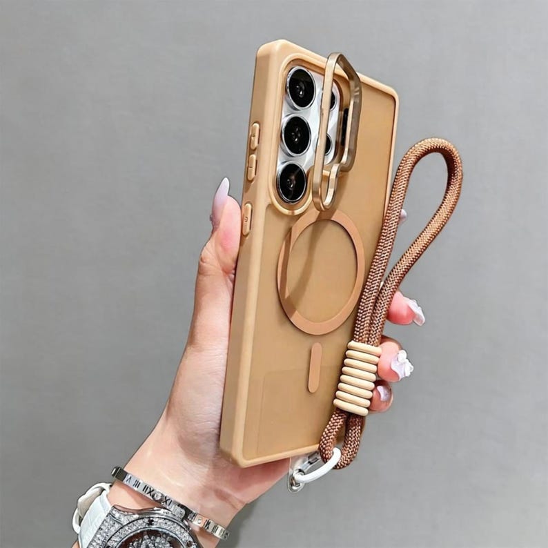 May include: A tan phone case with a gold ring holder and a matching brown lanyard. The case has a circular magnetic attachment and a vertical bar design. The lanyard features a woven cord and a series of beige beads. The phone case is held in a hand.