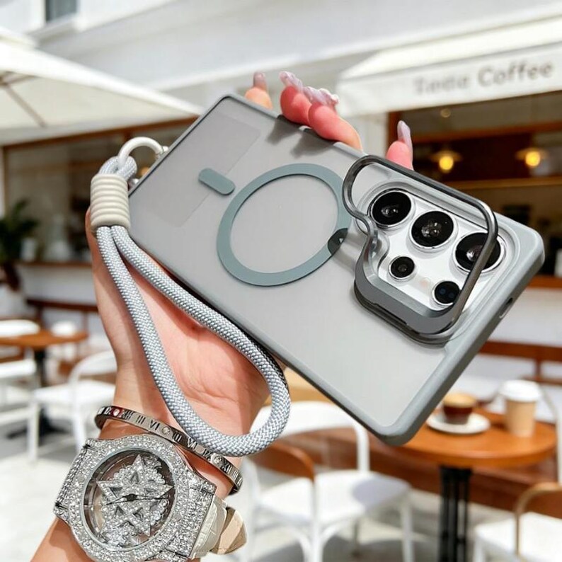 May include: A clear phone case with a gray border and a gray ring design. The case has a gray lanyard attached. The phone case has a silver camera protector. The image also shows a silver watch and bracelets.