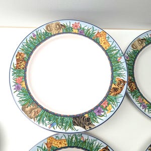 Op de afbeelding: Witte keramische borden met een jungle dierenontwerp. De borden hebben een blauwe rand met illustraties van olifanten, tijgers, zebra's en luipaarden te midden van groen gras en kleurrijke bloemen. De borden hebben een lichtroze midden.