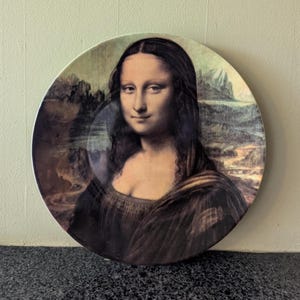 Peut inclure: Une assiette décorative ronde représentant la peinture de la Joconde. L'assiette présente le portrait emblématique d'une femme avec un sourire subtil, sur fond de paysage avec montagnes et ciel nuageux.