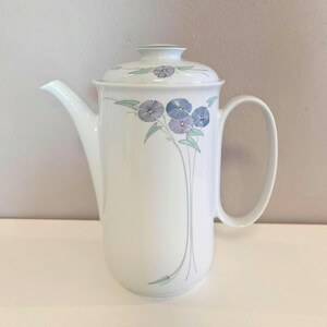 Könnte beinhalten: Weiße Keramik-Kaffeekanne mit Deckel, verziert mit einem floralen Muster in Blau- und Lilatönen. Die Kanne hat einen gebogenen Ausguss und einen großen Griff. Das florale Design befindet sich auf der Vorderseite und dem Deckel.