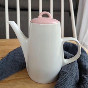 Może przedstawiać: Biały ceramiczny imbryk z różową pokrywką i uchwytem. Imbryk ma długi dzióbek i zakrzywiony uchwyt. Stoi na drewnianej powierzchni z niebieską tkaniną pod spodem.