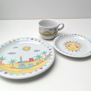 Op de afbeelding: Een witte keramische kinderservies set met een groot bord, een klein bord en een kopje. De borden en het kopje zijn versierd met een strandscène, inclusief een lachende zon, palmbomen en een kasteel. De set heeft een kleurrijk, speels ontwerp.