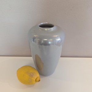 Könnte beinhalten: Eine hellgraue Keramikvase mit rundem Körper und schmalem Hals. Eine gelbe Zitrone liegt neben der Vase. Die Vase hat eine glänzende Oberfläche und reflektiert Licht. Die Vase ist ein dekorativer Gegenstand.