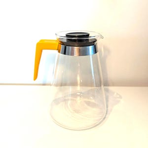 Può includere: Una caraffa di vetro trasparente con coperchio nero e manico giallo brillante. La caraffa ha una fascia argentata attorno alla parte superiore e un beccuccio per versare facilmente. Il design è moderno e minimalista.