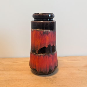 Może przedstawiać: Wazon z ceramiki w stylu vintage o uderzającym gradiencie kolorów. Góra i dół są ciemnobrązowe, przechodzące w żywą czerwień i czerń pośrodku. Wazon ma cylindryczny kształt z lekko rozszerzoną podstawą, idealny do dekoracji wnętrz.